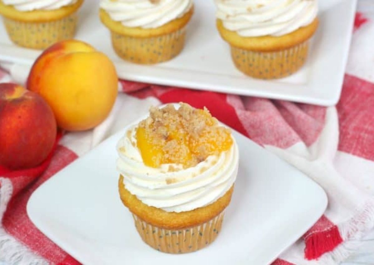 A vanilla cupcake with white frosting, peach slice, and crumb topping on a plate, with whole peaches and more cupcakes in the background.