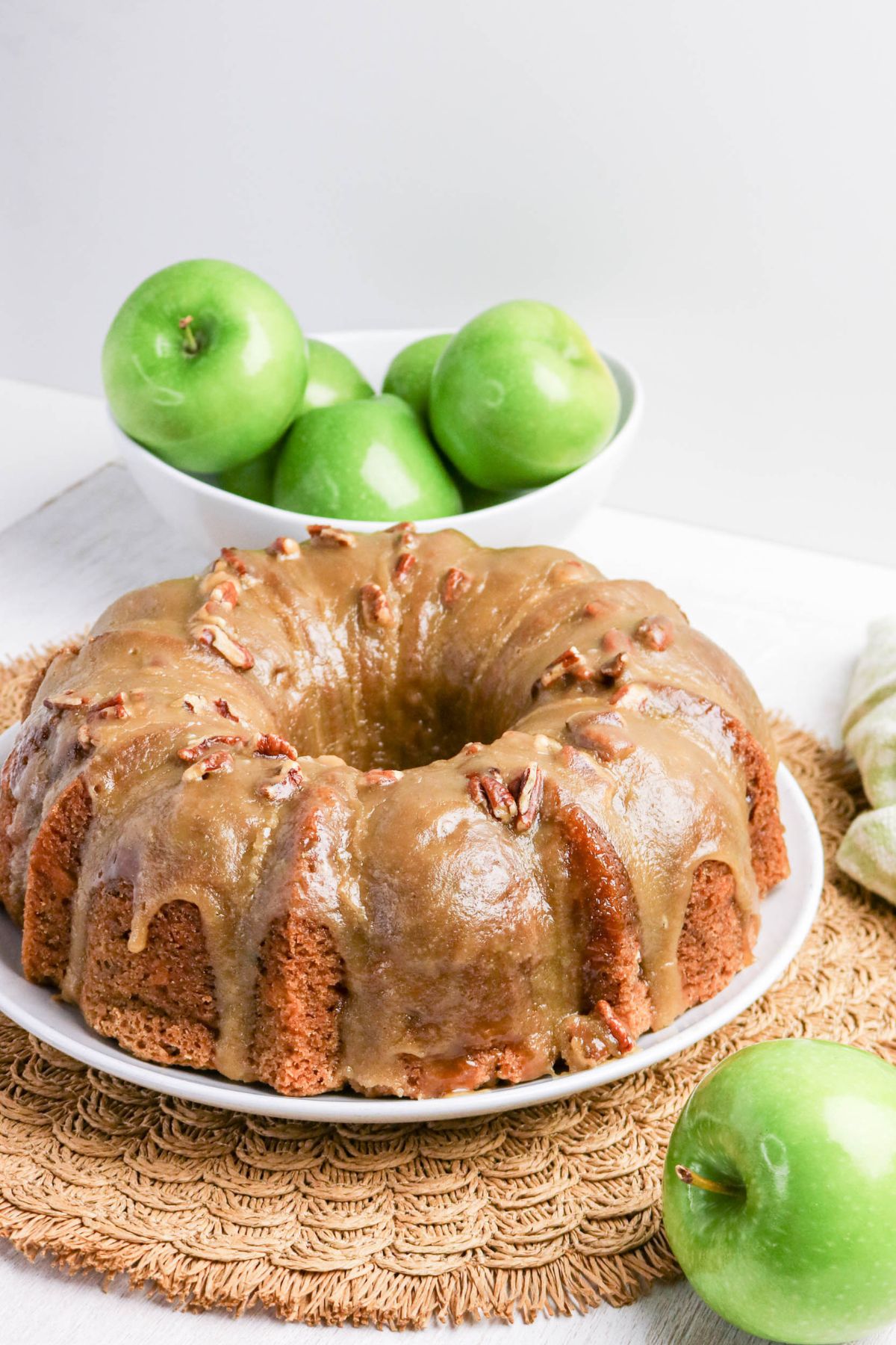 A bundt cake with glaze and pecans on a white plate sits on a woven mat, with a bowl of green apples and an apple in the foreground.