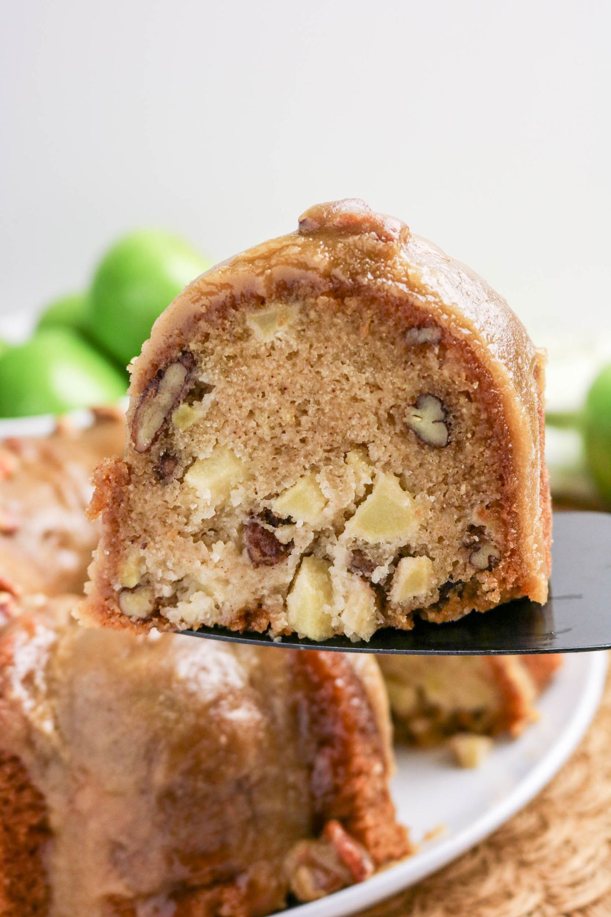 A close-up of a slice of apple pecan bundt cake with chunks of apple and pecans, topped with a caramel glaze, held above a whole cake.