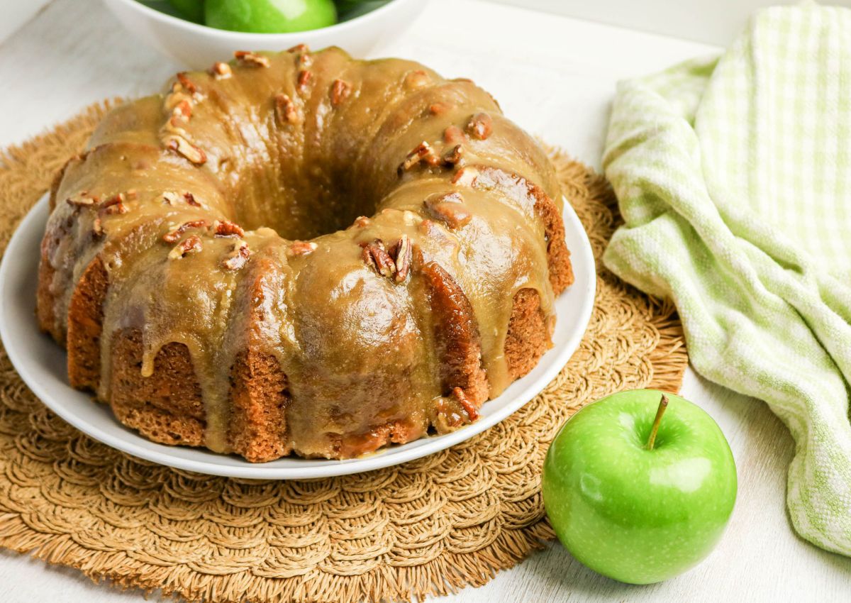 A caramel-glazed bundt cake with pecans sits on a white plate next to a green apple and a green checkered cloth. A bowl of green apples is in the background.