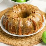 A bundt cake with caramel glaze and pecans sits on a white plate atop a woven placemat, with green apples in the background.