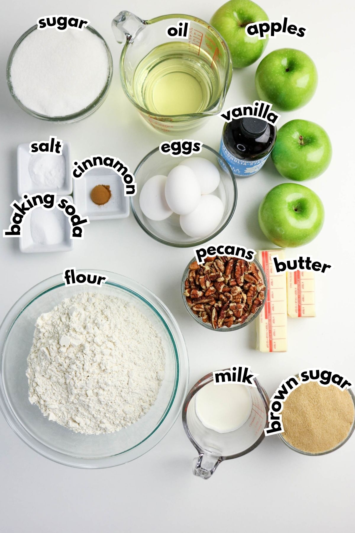 Baking ingredients arranged on a white surface, including flour, sugar, brown sugar, eggs, oil, butter, milk, pecans, apples, vanilla, baking soda, salt, and cinnamon.