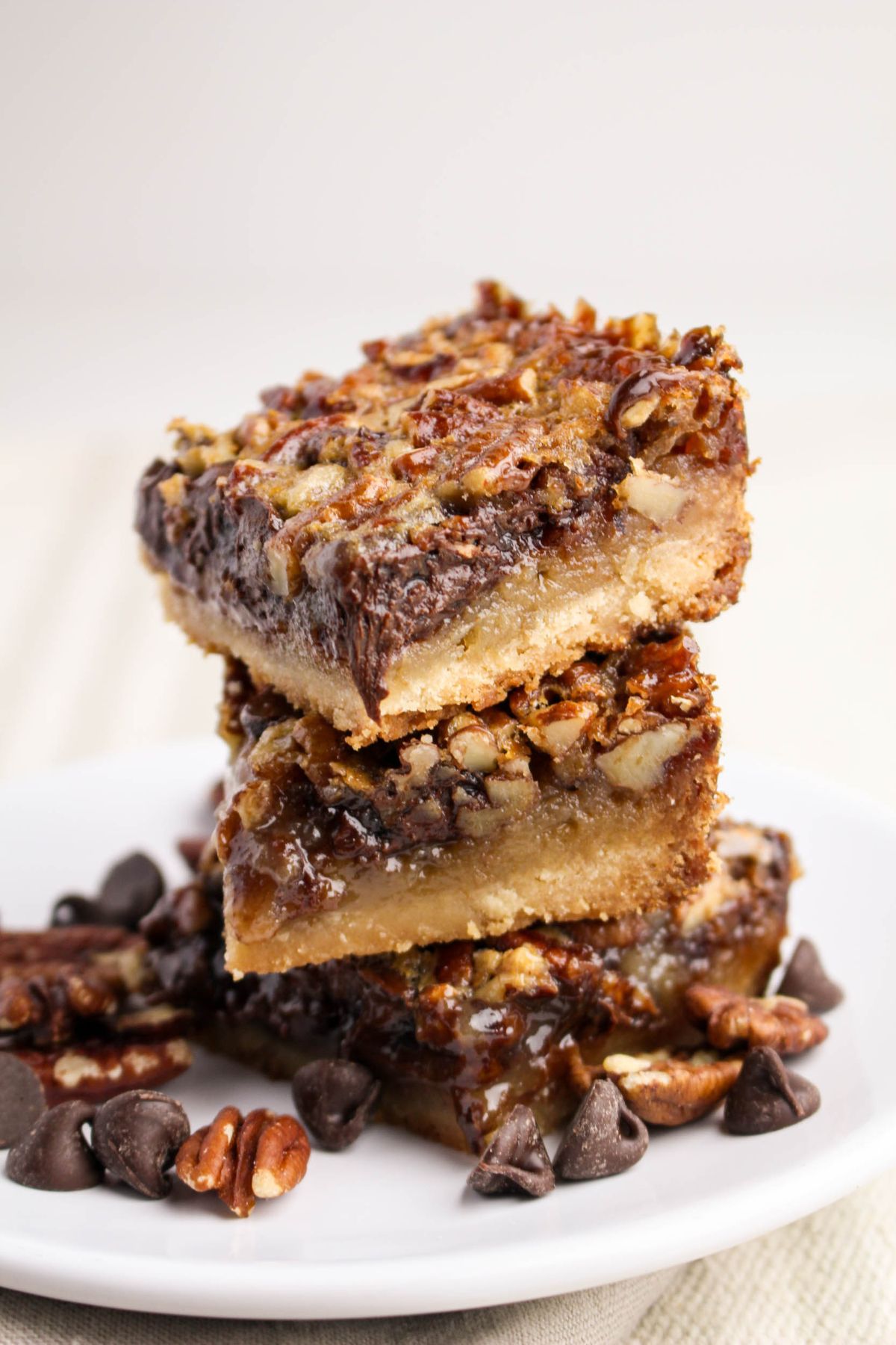 Three stacked pecan bars with a gooey filling sit on a white plate, surrounded by chocolate chips and pecan pieces.