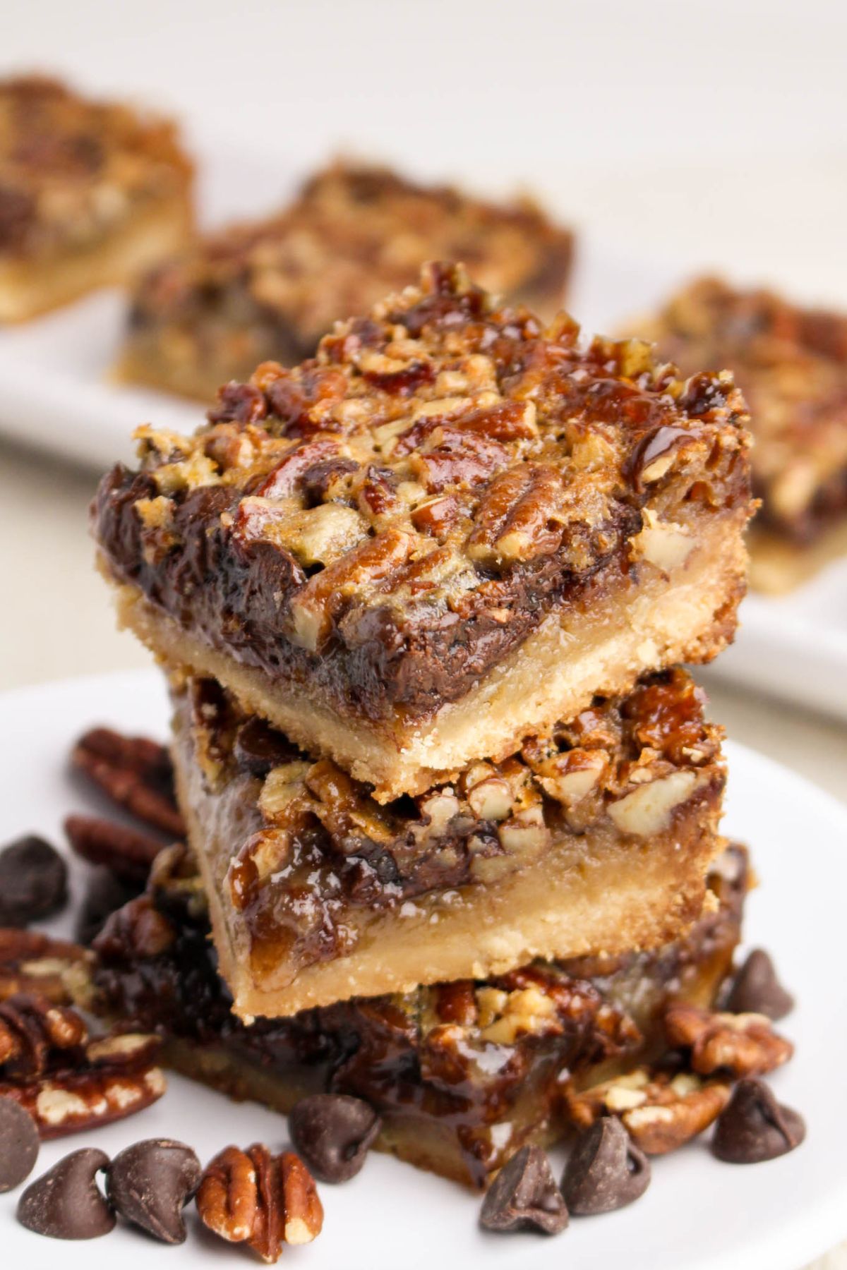 Two pecan pie bars are stacked on a plate, surrounded by pecans and chocolate chips, with more bars visible in the background.