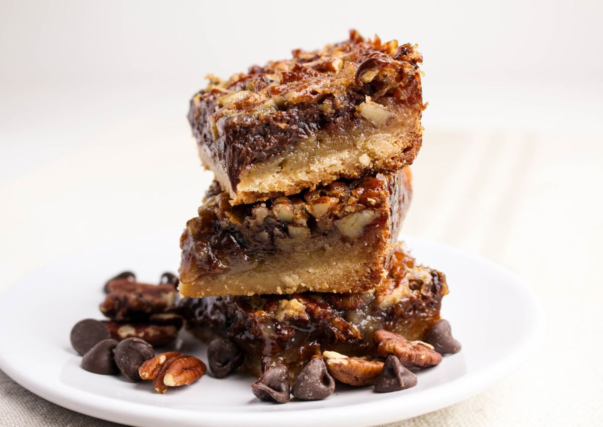Three pecan bars stacked on a white plate, surrounded by chocolate chips and pecan pieces, with a white background.