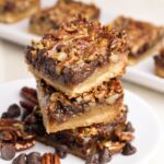 Two stacked pecan pie bars on a white plate, surrounded by pecan halves and chocolate chips, with more bars visible in the background.