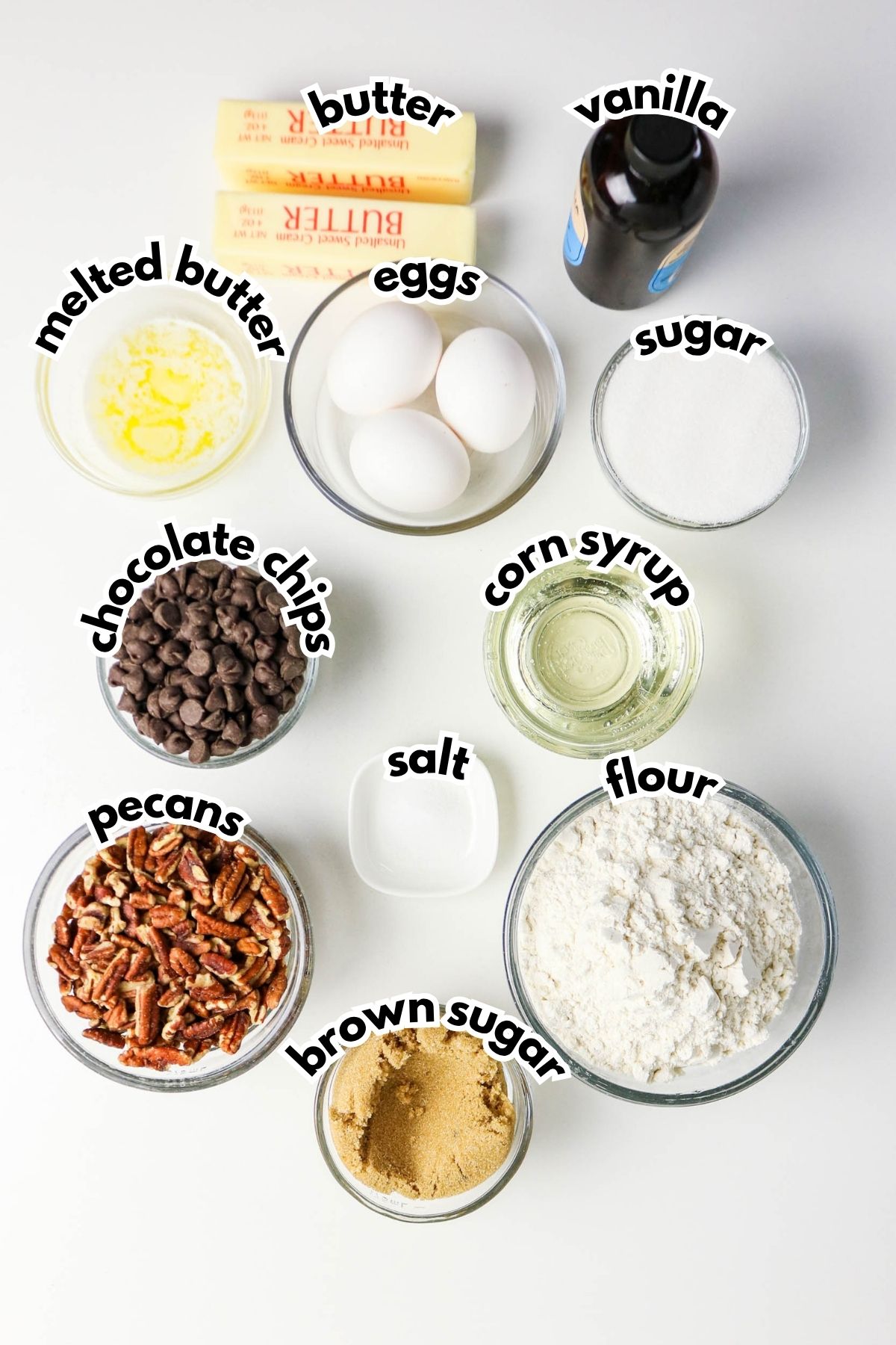 Various labeled baking ingredients are arranged on a white surface, including butter, eggs, vanilla, sugar, corn syrup, flour, brown sugar, salt, pecans, chocolate chips, and melted butter.