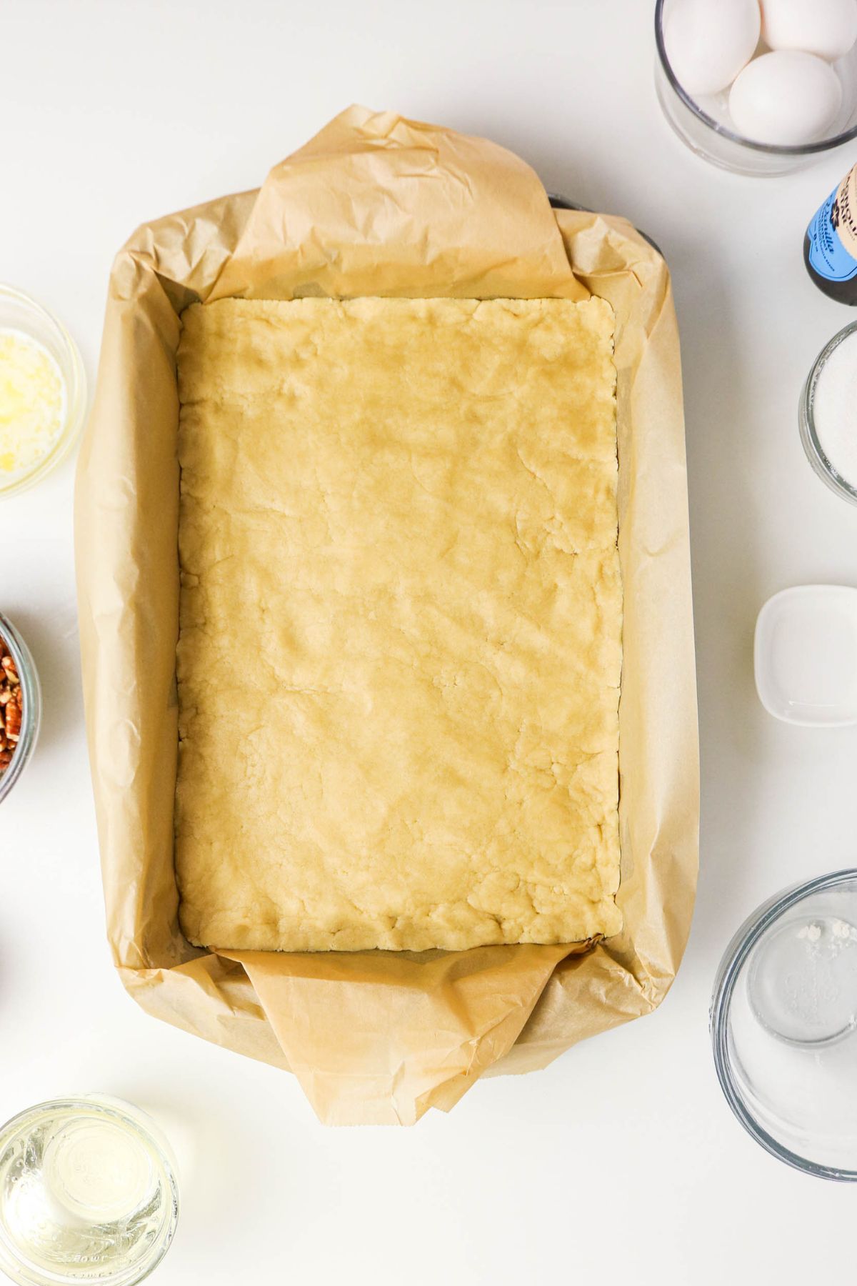 Unbaked dough pressed into a parchment-lined rectangular baking pan, surrounded by bowls containing eggs and baking ingredients.
