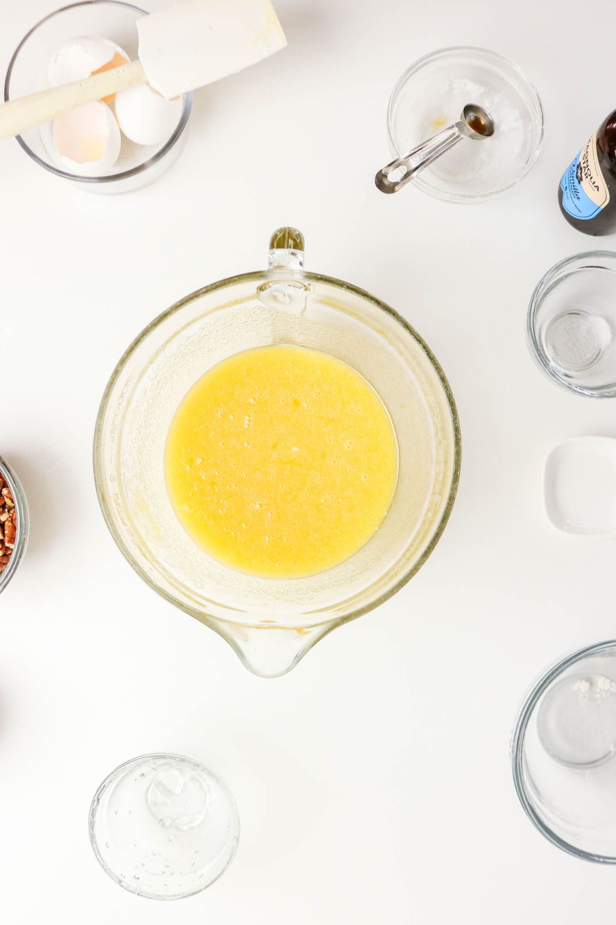 A glass bowl with a yellow liquid mixture sits on a white surface, surrounded by bowls of baking ingredients, a spatula, and measuring spoons.