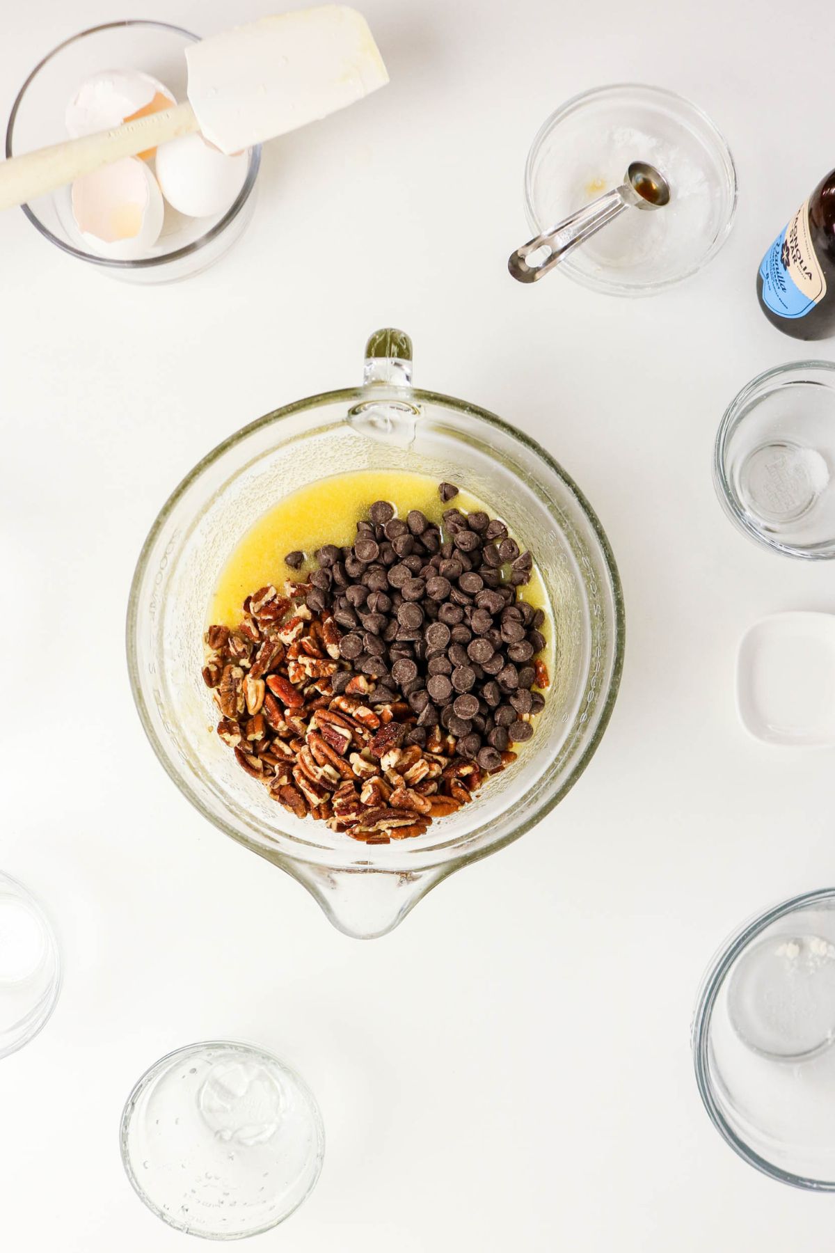 A glass mixing bowl with chopped pecans and chocolate chips on top of melted butter, surrounded by empty bowls, cracked eggs, a spatula, and a bottle of vanilla extract on a white surface.