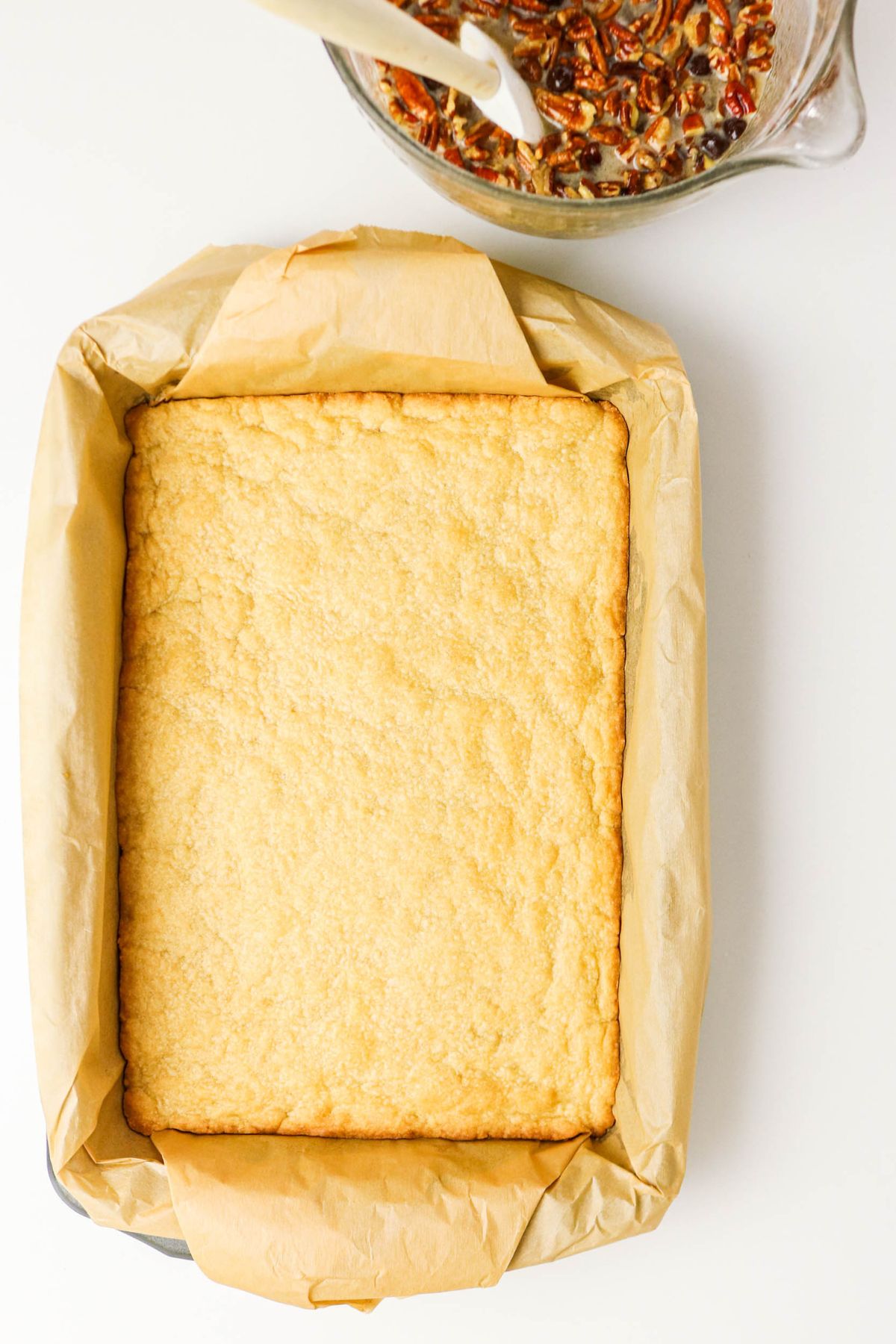 A rectangular baking pan lined with parchment paper contains a baked crust. Next to it, a glass bowl holds a pecan mixture with a spatula.