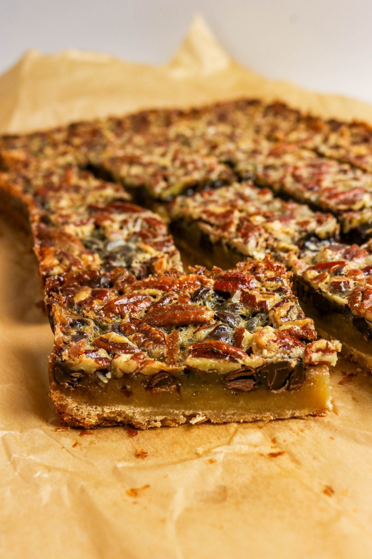 A close-up of sliced pecan pie bars on parchment paper, showing a golden crust, gooey filling, and pecan topping.
