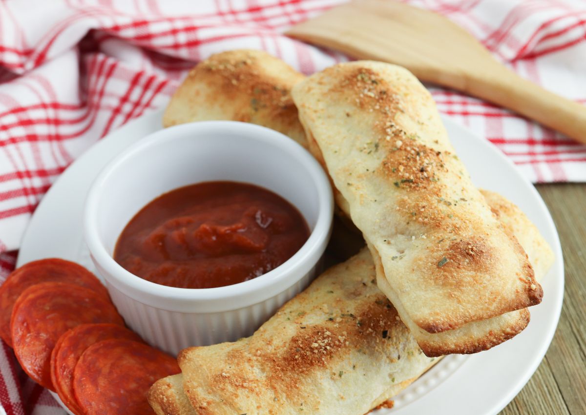 Three baked breadsticks on a plate with a side of marinara sauce in a ramekin and slices of pepperoni, set on a red and white checkered cloth.