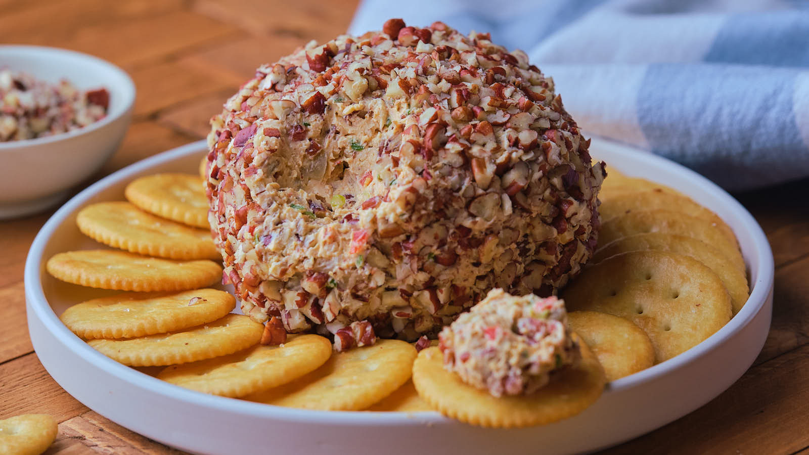 A cheese ball coated in chopped nuts is served on a white plate with round crackers arranged around it.