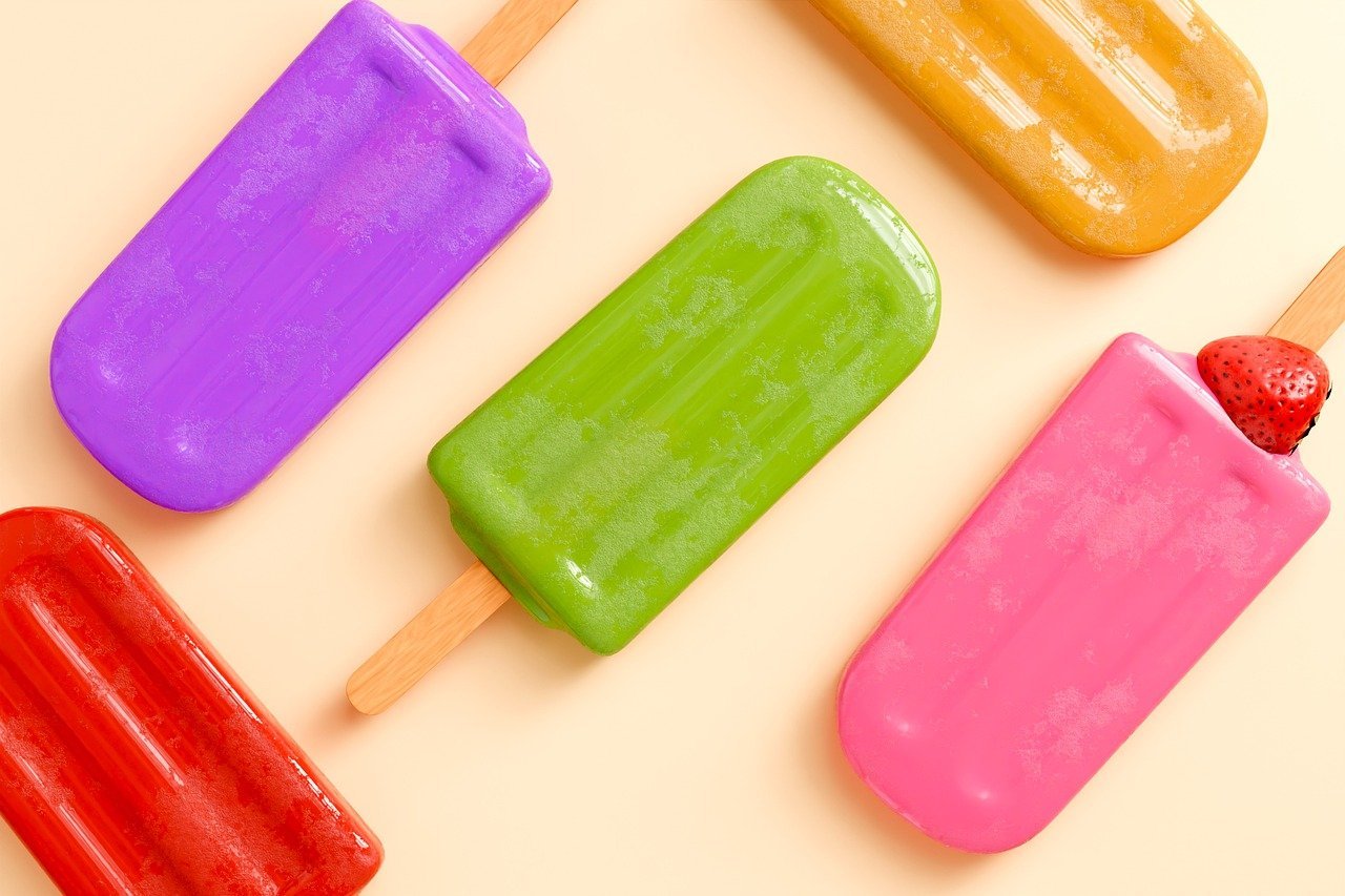 Five colorful popsicles—purple, green, yellow, red, and pink—are arranged on a light background, with a strawberry partly visible near the pink popsicle.