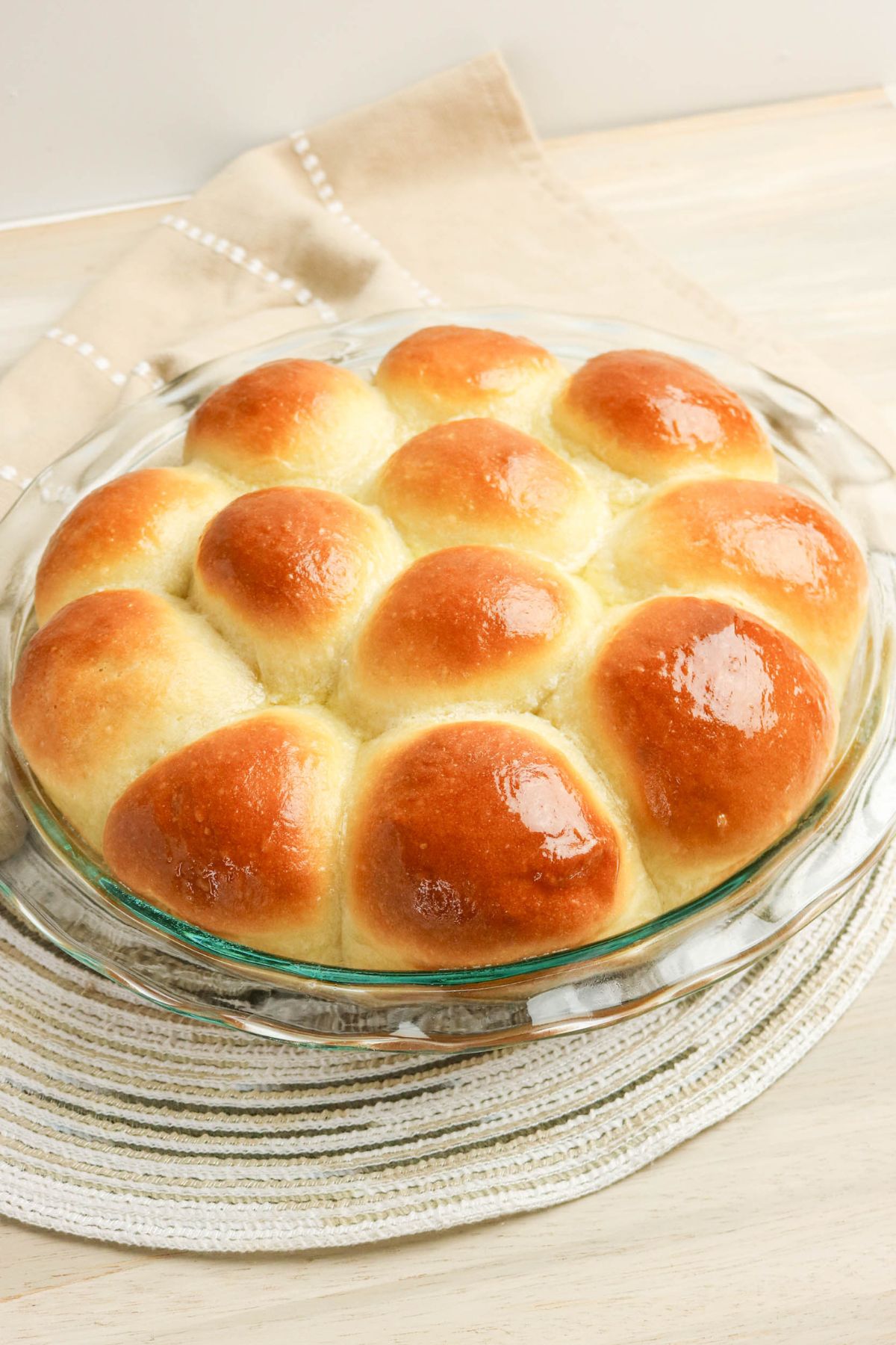 A glass pie dish filled with twelve golden-brown, shiny dinner rolls sits on a white woven placemat and a beige napkin.