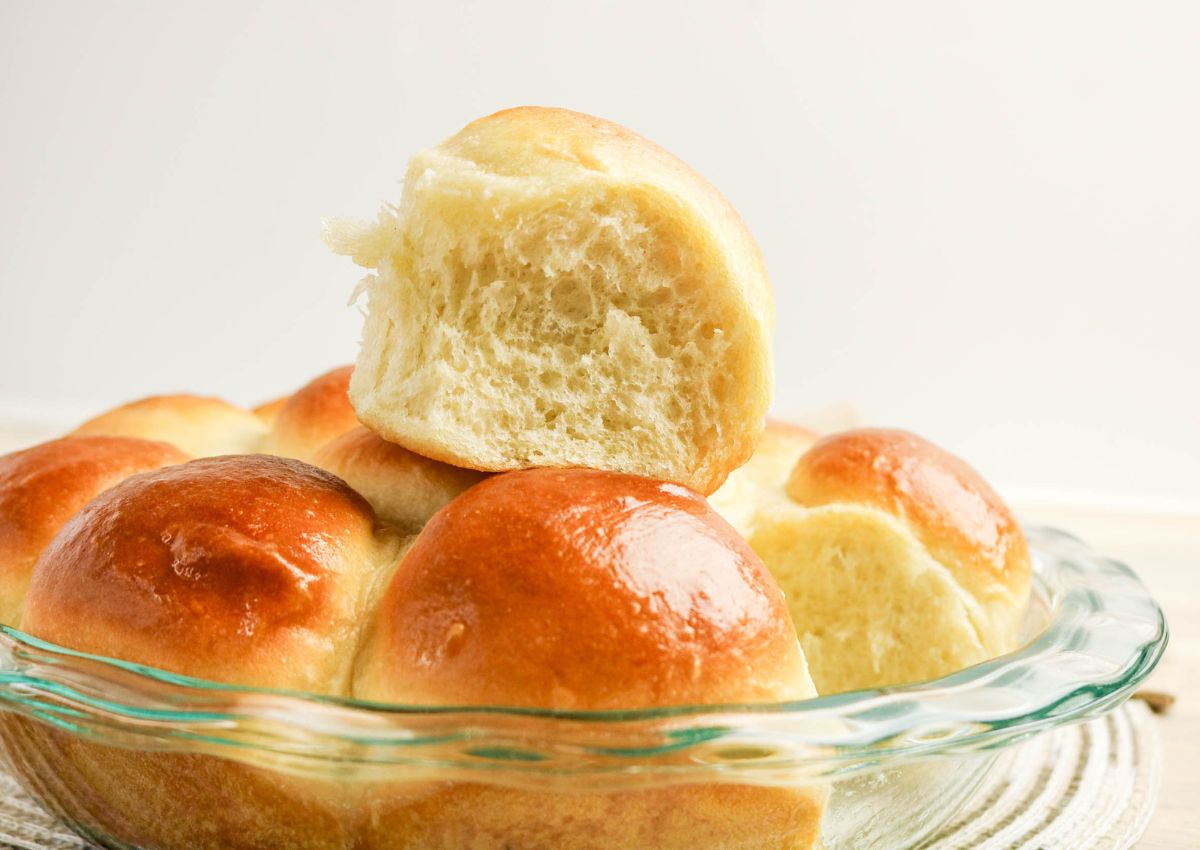A glass dish filled with golden brown, shiny dinner rolls, with one roll pulled apart to show its soft, fluffy interior.