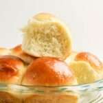 A glass dish filled with soft, golden-brown dinner rolls; one roll is broken open and placed on top, showing its fluffy interior.