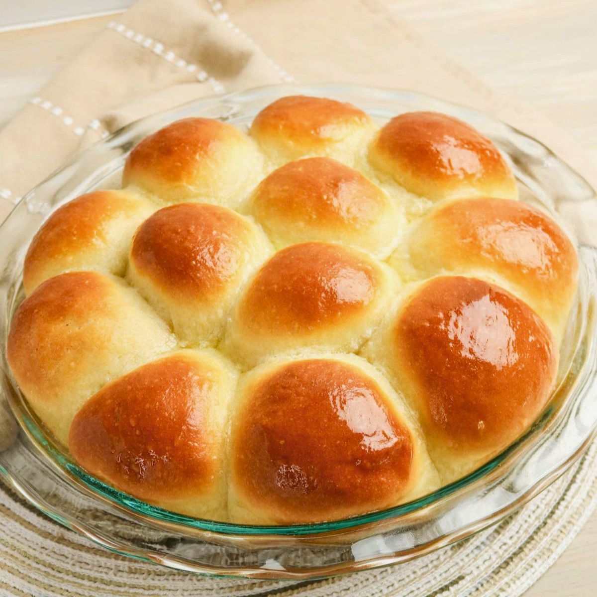 A round glass dish filled with twelve golden brown, shiny pull apart rolls arranged in a circle on a beige cloth.