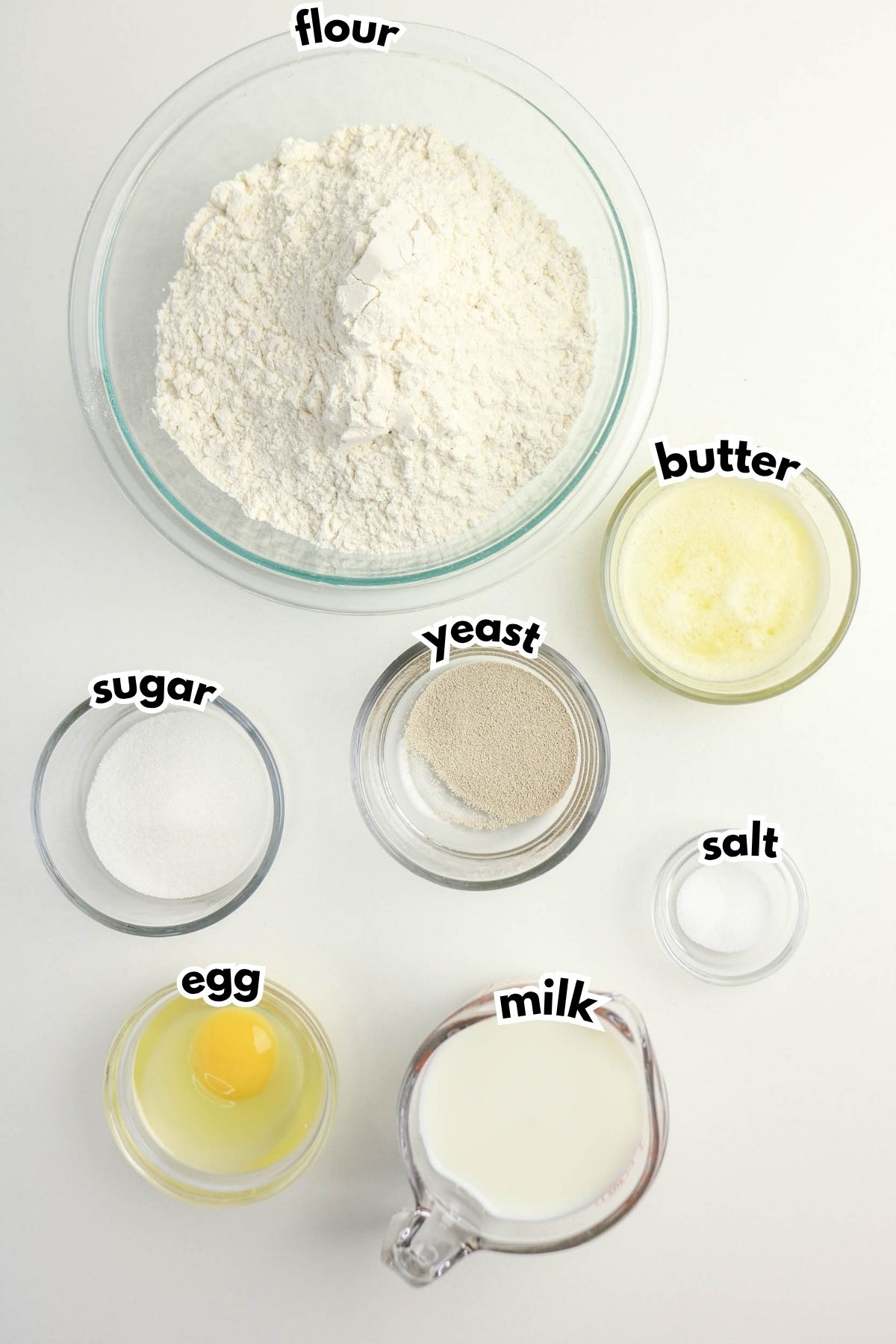 Seven labeled ingredients for bakingโflour, butter, sugar, yeast, salt, egg, and milkโare shown in separate bowls and a measuring cup on a white surface.