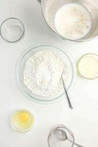 Bowls containing flour, sugar, and salt, an egg, melted butter, milk with yeast, and water are arranged on a white surface, viewed from above.
