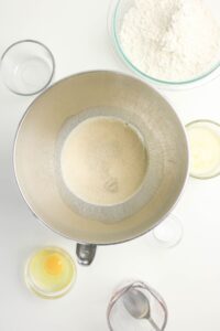 A mixing bowl with yeast dissolving in water, surrounded by bowls containing flour, melted butter, an egg, and measuring cups on a white surface.