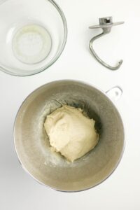 A metal mixing bowl with dough, an empty glass bowl, and a dough hook attachment on a white surface.