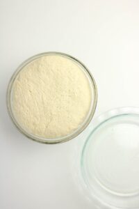 A glass bowl filled with risen dough sits next to an empty glass lid on a white surface.