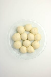 A glass dish containing twelve evenly spaced, round dough balls on a white surface.