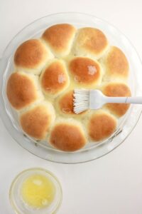 A glass dish of golden brown dinner rolls is being brushed with melted butter, with a bowl of butter nearby on a white surface.