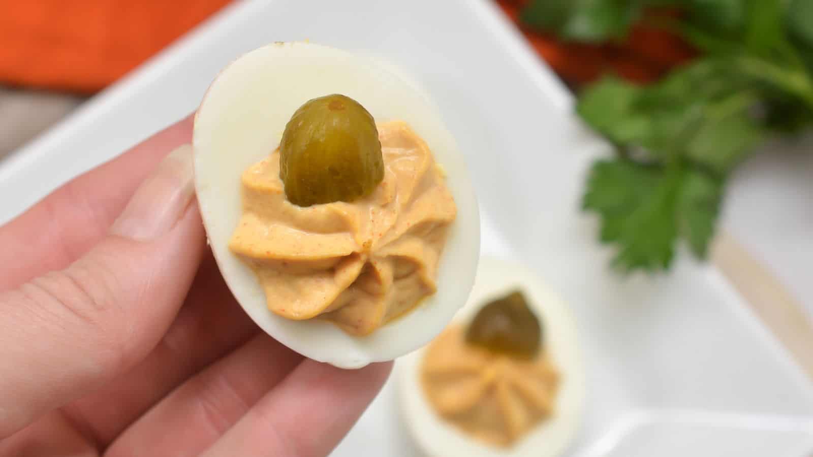 A hand holds a deviled egg topped with a dollop of creamy filling and a small slice of pickle; another deviled egg is on a plate in the background.