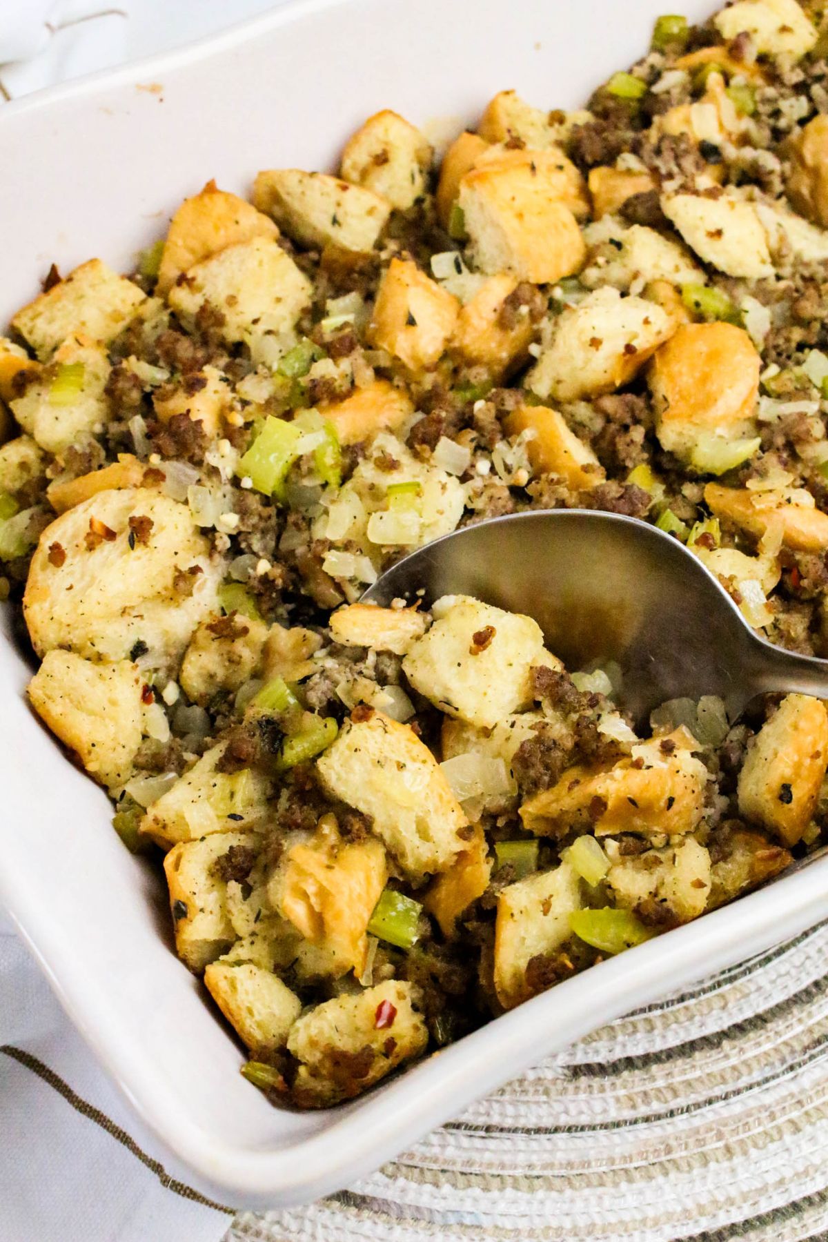 A baking dish filled with bread stuffing mixed with sausage, celery, and onions, with a serving spoon resting inside.