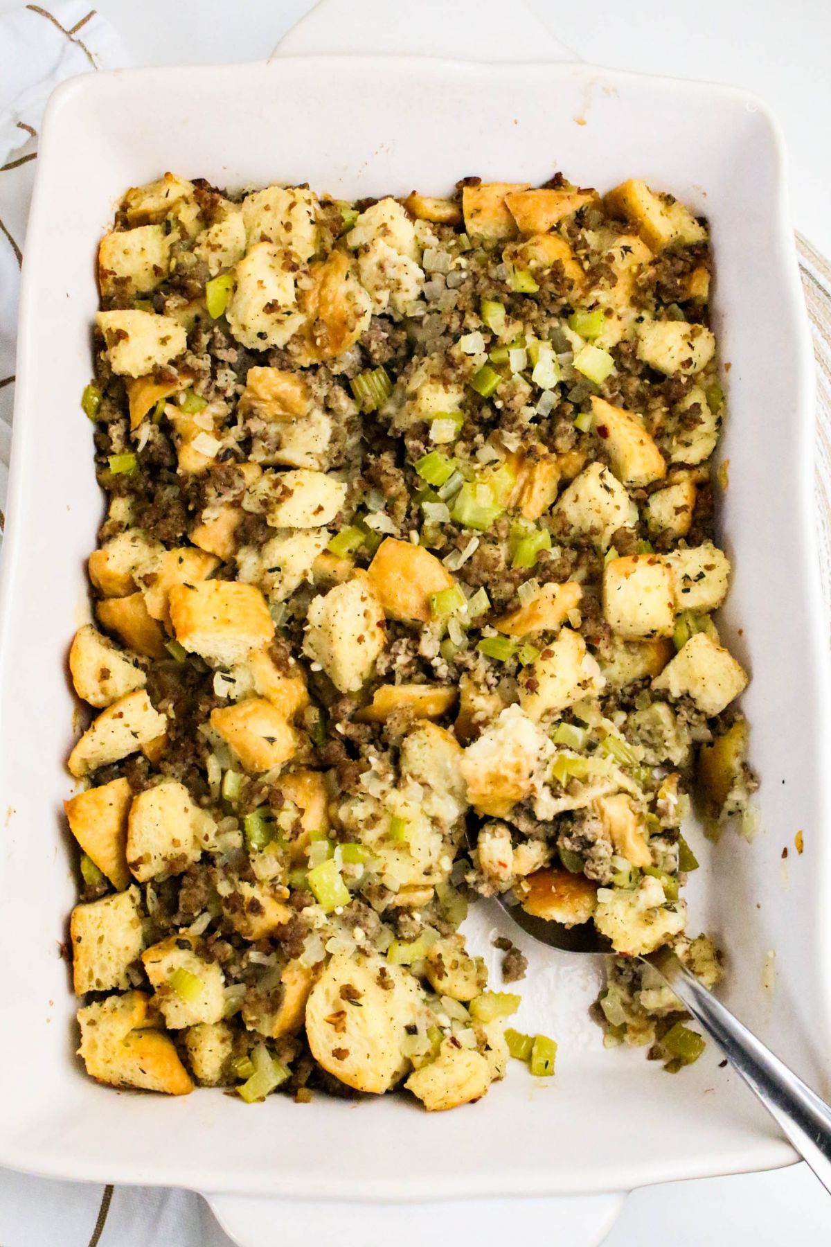 A rectangular white baking dish filled with bread stuffing mixed with celery and herbs; a serving spoon is placed in the dish.