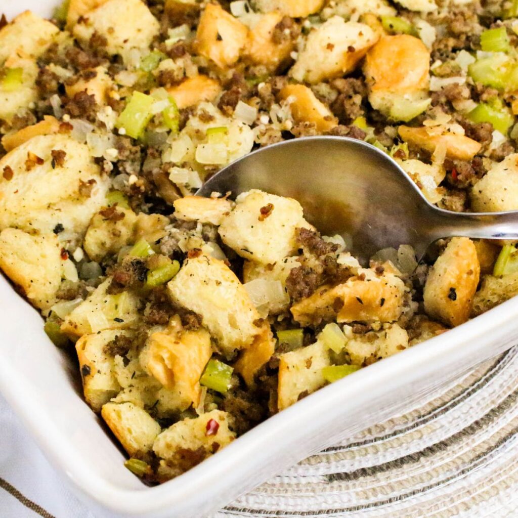 A close-up of a baking dish filled with bread stuffing containing chunks of bread, ground meat, celery, and onions, with a serving spoon resting in the dish.
