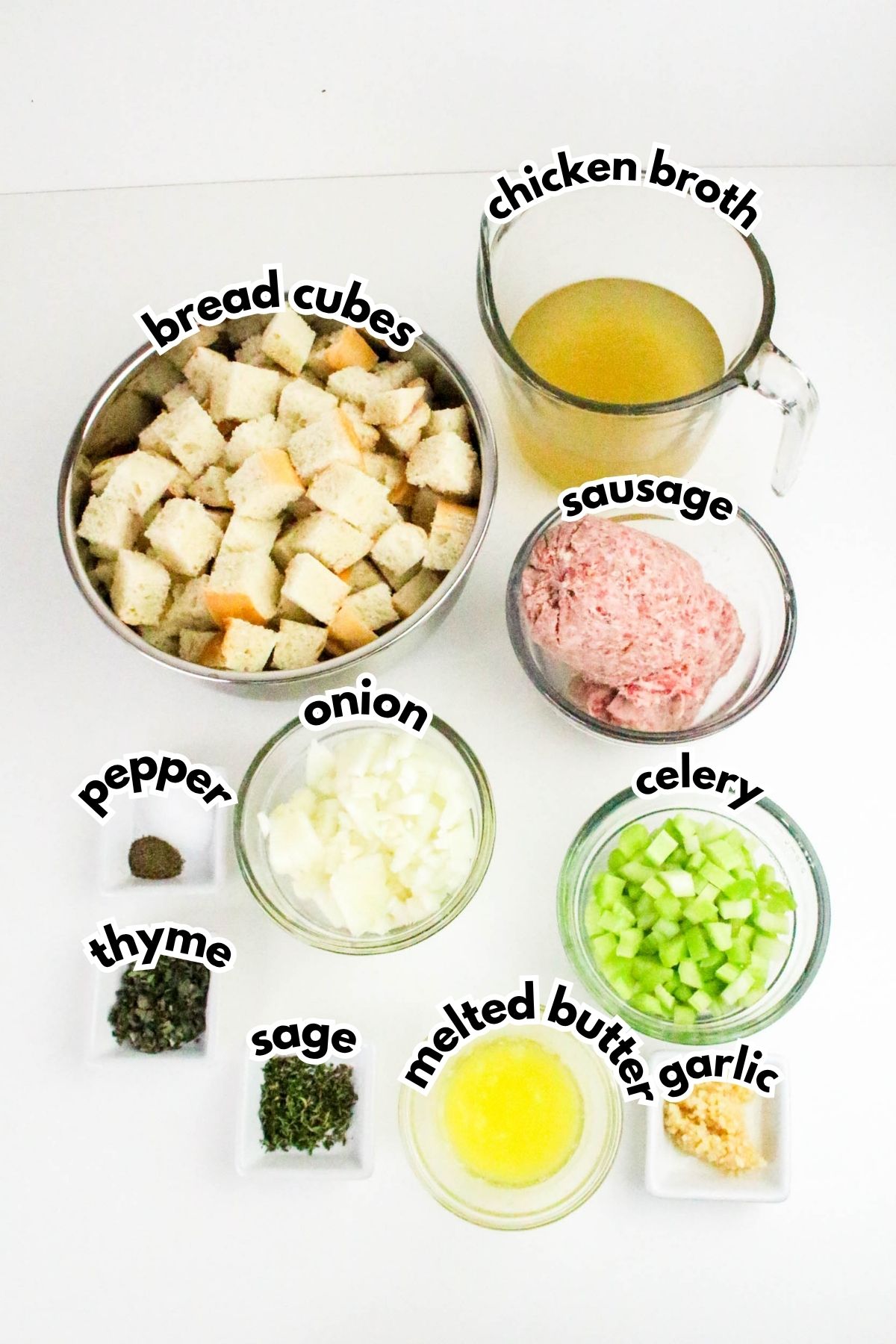 Ingredients for stuffing recipe arranged on a white surface: bread cubes, chicken broth, sausage, diced onion, chopped celery, melted butter, minced garlic, sage, thyme, and pepper.