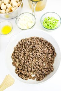 Cooked ground sausage in a white skillet, surrounded by bowls of cubed bread, melted butter, chopped onion, celery, and broth on a white surface.