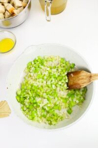 A white pan with chopped celery and onions being sautรฉed, stirred with a wooden spoon. Bread cubes, melted butter, and broth are visible on the side.