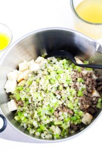 A mixing bowl containing cubed bread, cooked ground meat, chopped celery, and sautรฉed onions, with a black spoon inside; cups of broth and melted butter nearby.