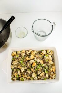 A white baking dish filled with stuffing mixture containing bread cubes and vegetables sits on a white surface next to a mixing bowl, a small glass bowl, and a glass measuring cup.