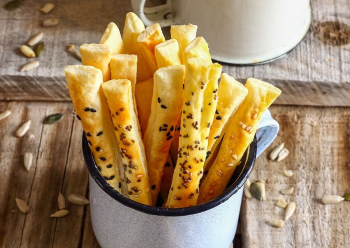 A white enamel mug holds several golden-brown breadsticks topped with seeds, sitting on a rustic wooden surface with scattered seeds around.