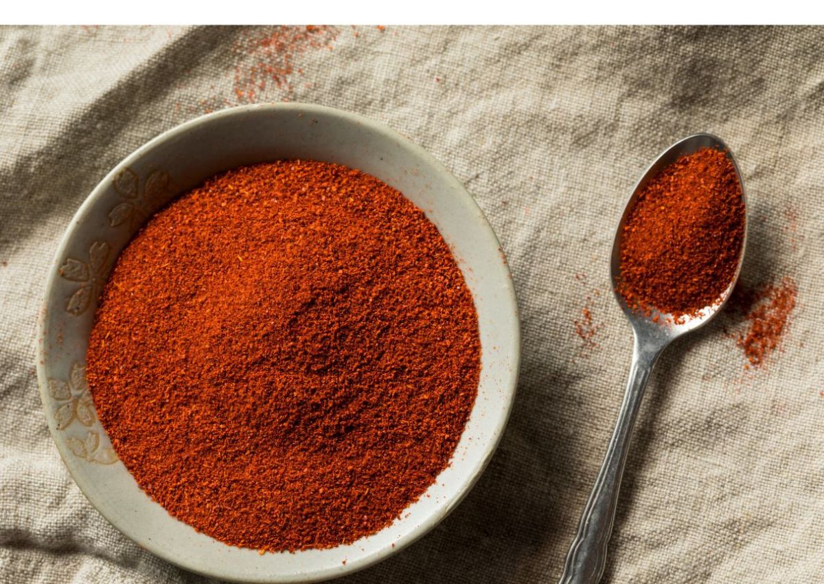 A bowl and a spoon filled with ground red spice, likely paprika or chili powder, on a textured cloth surface.
