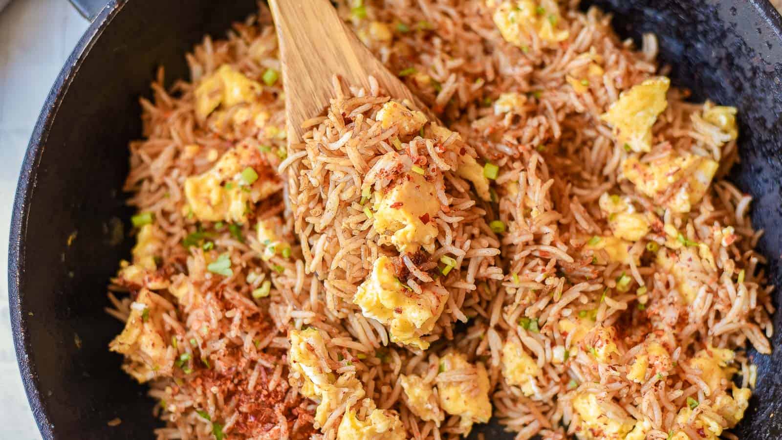 A pan of Spicy Egg Fried Rice with scrambled eggs, garnished with seasonings and stirred with a wooden spatula.
