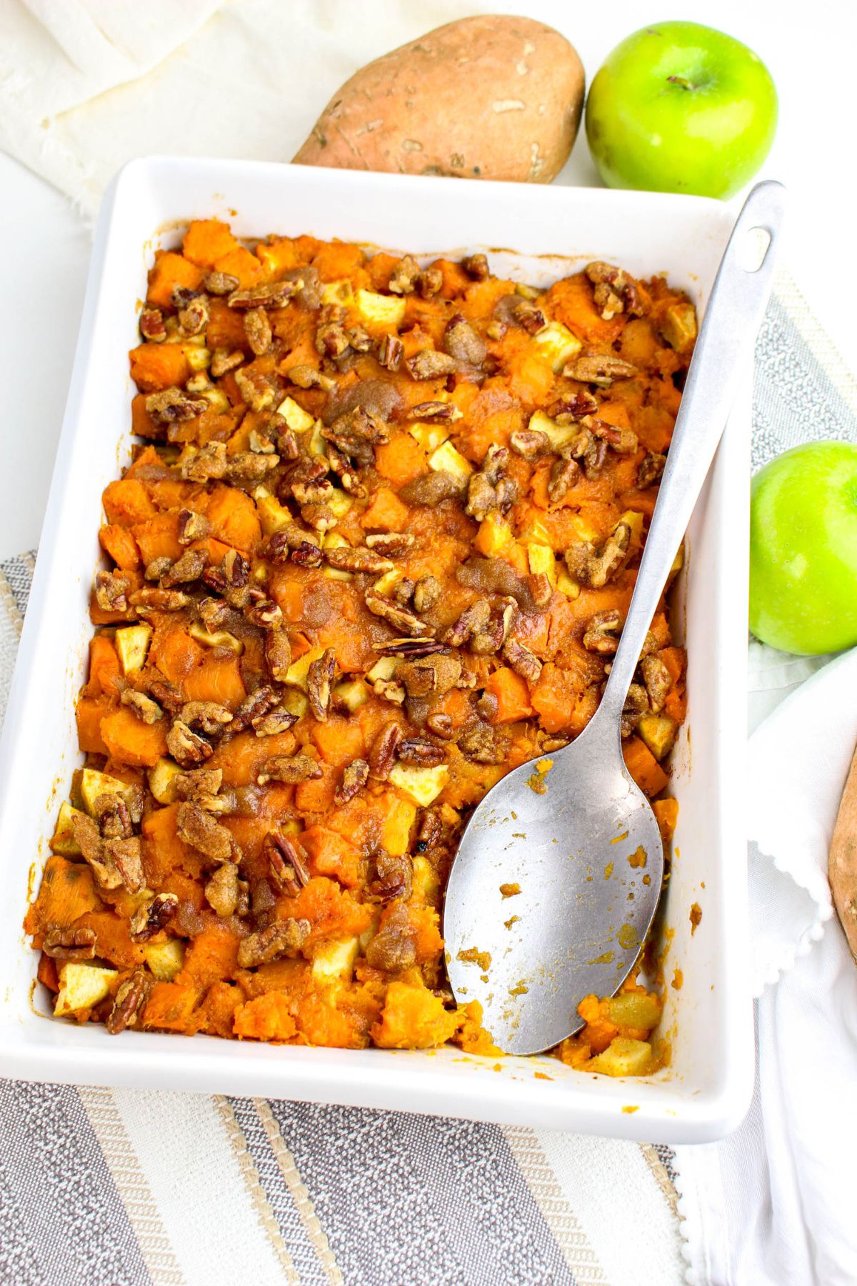 A baked casserole dish filled with sweet potato and apple chunks topped with pecans, with a large serving spoon resting inside. Whole sweet potatoes and green apples are nearby.