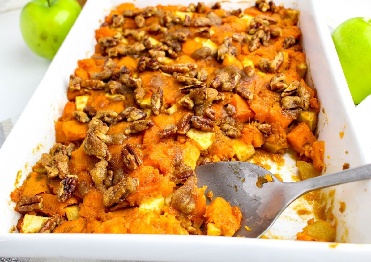 A white baking dish filled with sweet potato casserole topped with pecans, partially served with a metal spoon; green apples are in the background.