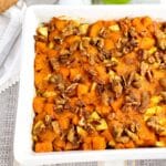 A baked sweet potato casserole topped with chopped pecans, served in a white rectangular dish on a patterned placemat.