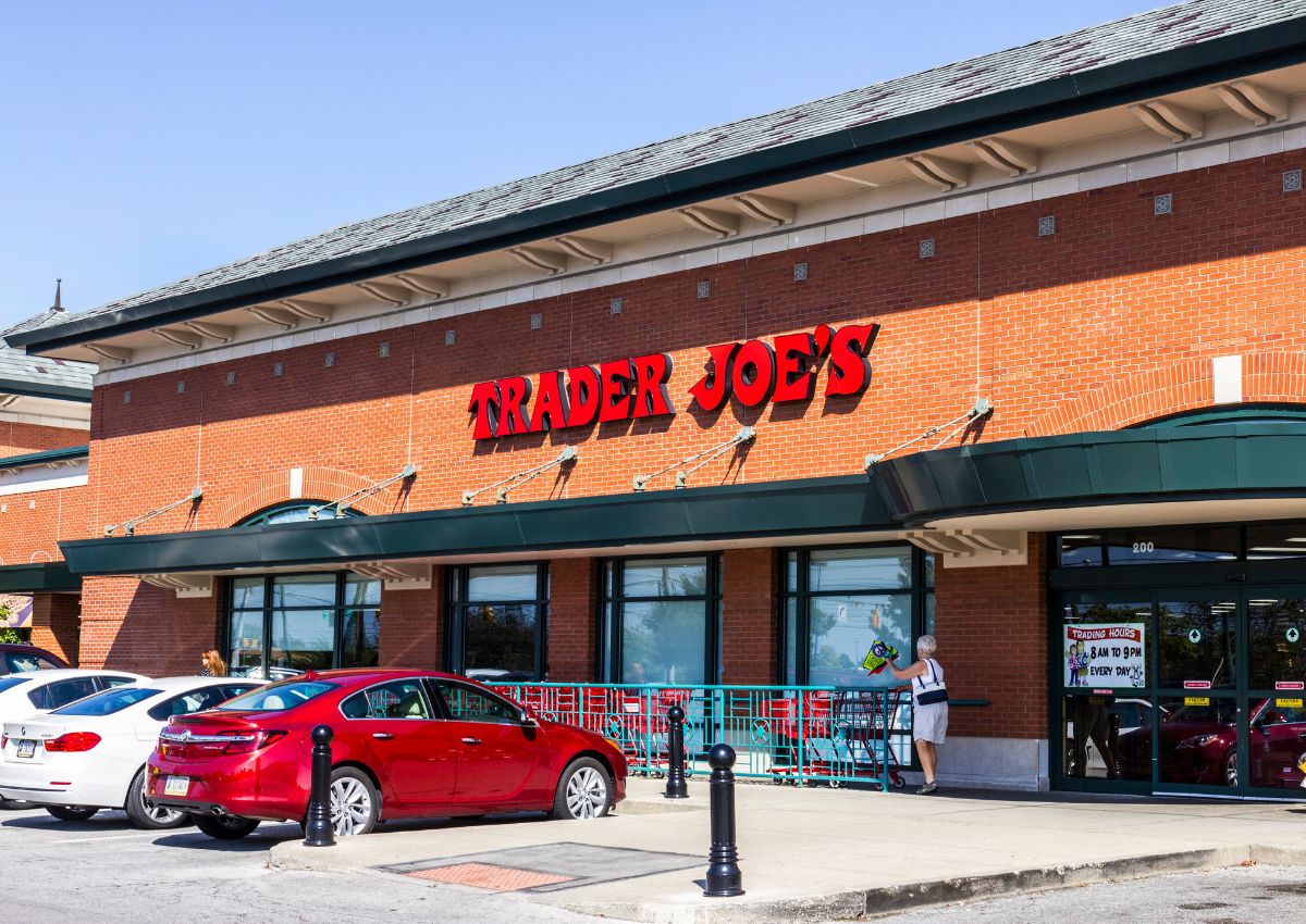 The exterior of a Trader Joe’s grocery store with parked cars and a person entering the building on a sunny day.