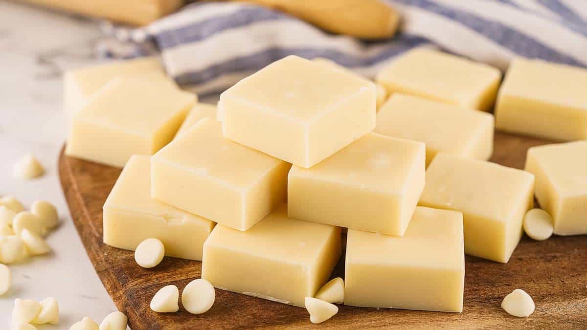 White chocolate fudge squares are stacked on a wooden board, with white chocolate chips scattered around and a striped cloth in the background.