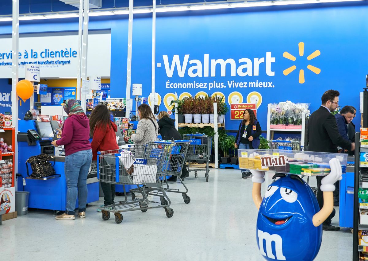 Customers with shopping carts wait in line at a Walmart checkout counter; a blue M&M character display stands nearby. Signs are in French and English.