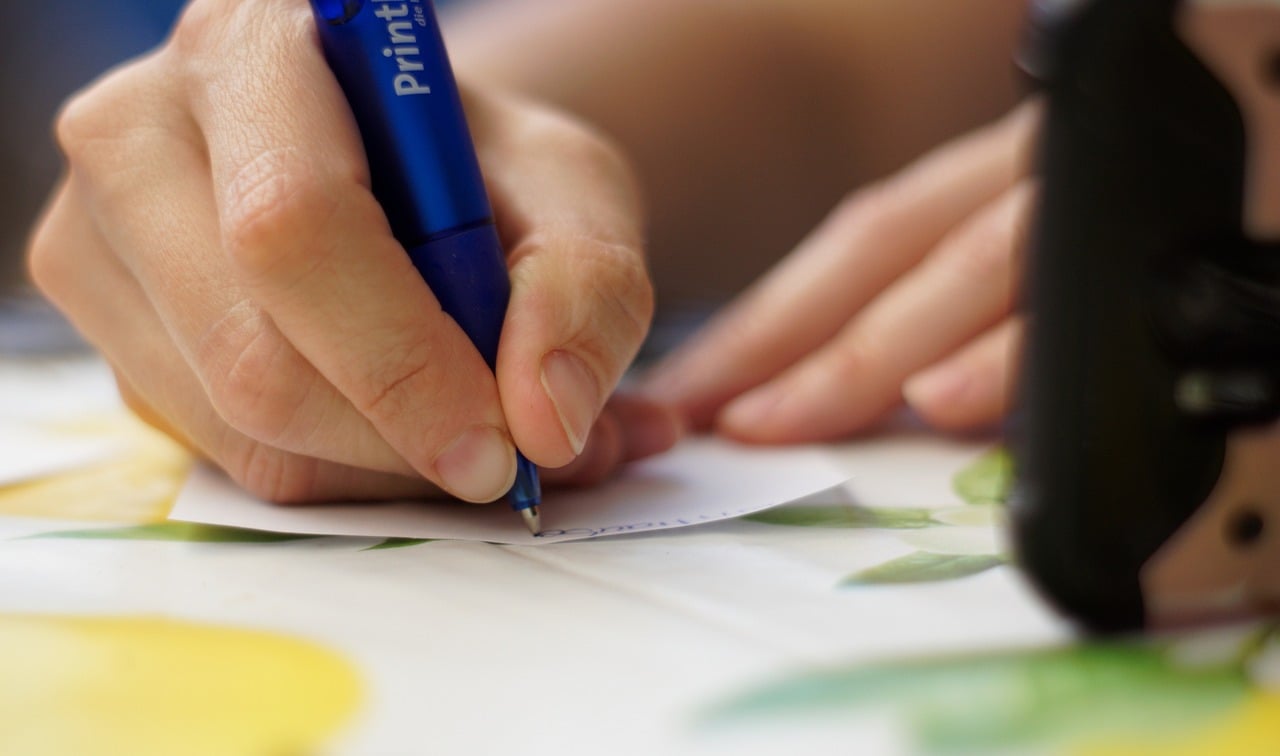 A person writes on a small piece of paper with a blue pen while resting their hand on a table with a lemon-patterned tablecloth.