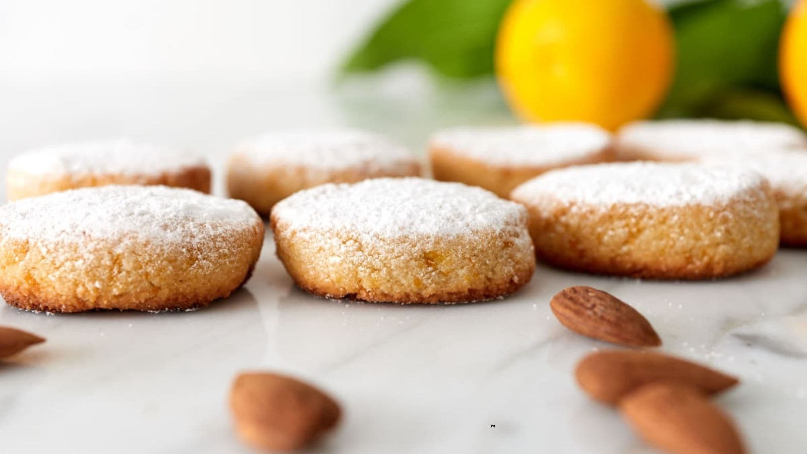 Round almond cookies dusted with powdered sugar are arranged on a marble surface with whole almonds and lemons in the background.